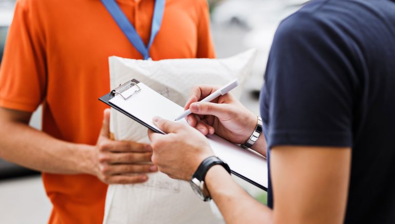 A person wearing an orange T-shirt and a name tag is delivering a parcel to a client, who is putting his signature on the receipt. Friendly worker, high quality delivery service.