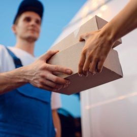 Deliveryman in uniform gives parcel to female recipient at the car, delivery service. Man holding cardboard package near the vehicle, male deliver and woman, courier or shipping job