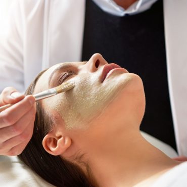 Unrecognizable male beautician putting mask on female face lying with eyes closed with brush in salon.