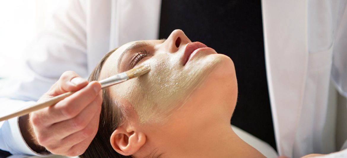 Unrecognizable male beautician putting mask on female face lying with eyes closed with brush in salon.