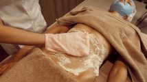 Female masseur rubbing cream on stomach of young slim woman in towel, professional massage. Massaging and relaxation, body and skin care. Attractive lady in spa salon